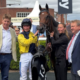 Jockey in yellow silks holding the reins of a dark horse, flanked by smiling spectators in smart casual attire at a racecourse with tents and banners in the background.