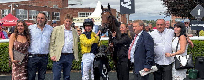 Jockey in yellow silks holding the reins of a dark horse, flanked by smiling spectators in smart casual attire at a racecourse with tents and banners in the background.