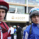 Two jockeys in red-and-white and blue racing silks with helmets stand together at Ascot racecourse, with spectators and an ASCOT sign in the background.