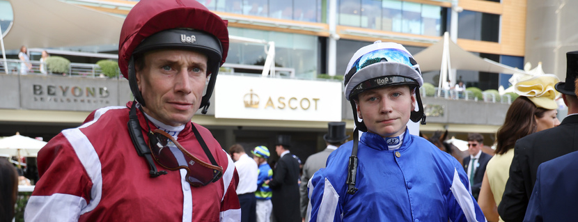 Two jockeys in red-and-white and blue racing silks with helmets stand together at Ascot racecourse, with spectators and an ASCOT sign in the background.